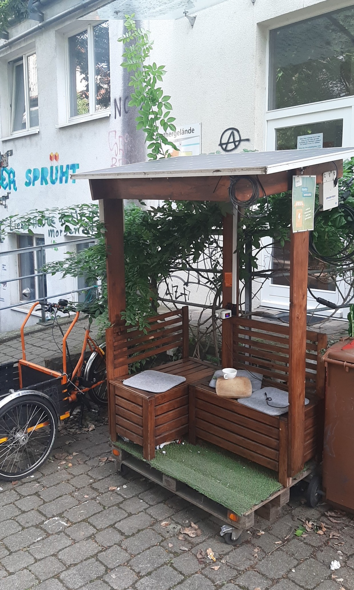 Bild von der solartankstelle auf dem grether Gelände beim cccfr und strandcafe,
auf holzpallette gebaute Bänke mit Dach aus grossen Solarpanel.
Daneben ein orangenes lastendreirad von lastenvelo Freiburg an seinem Parkplatz, wird gerade geladen.